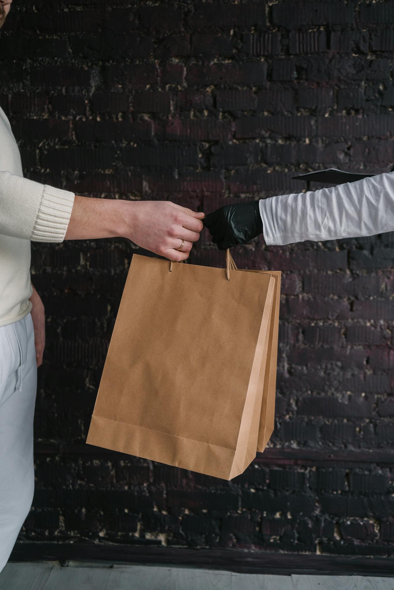 Close-up of two hands exchanging a brown paper shopping bag against a dark background.