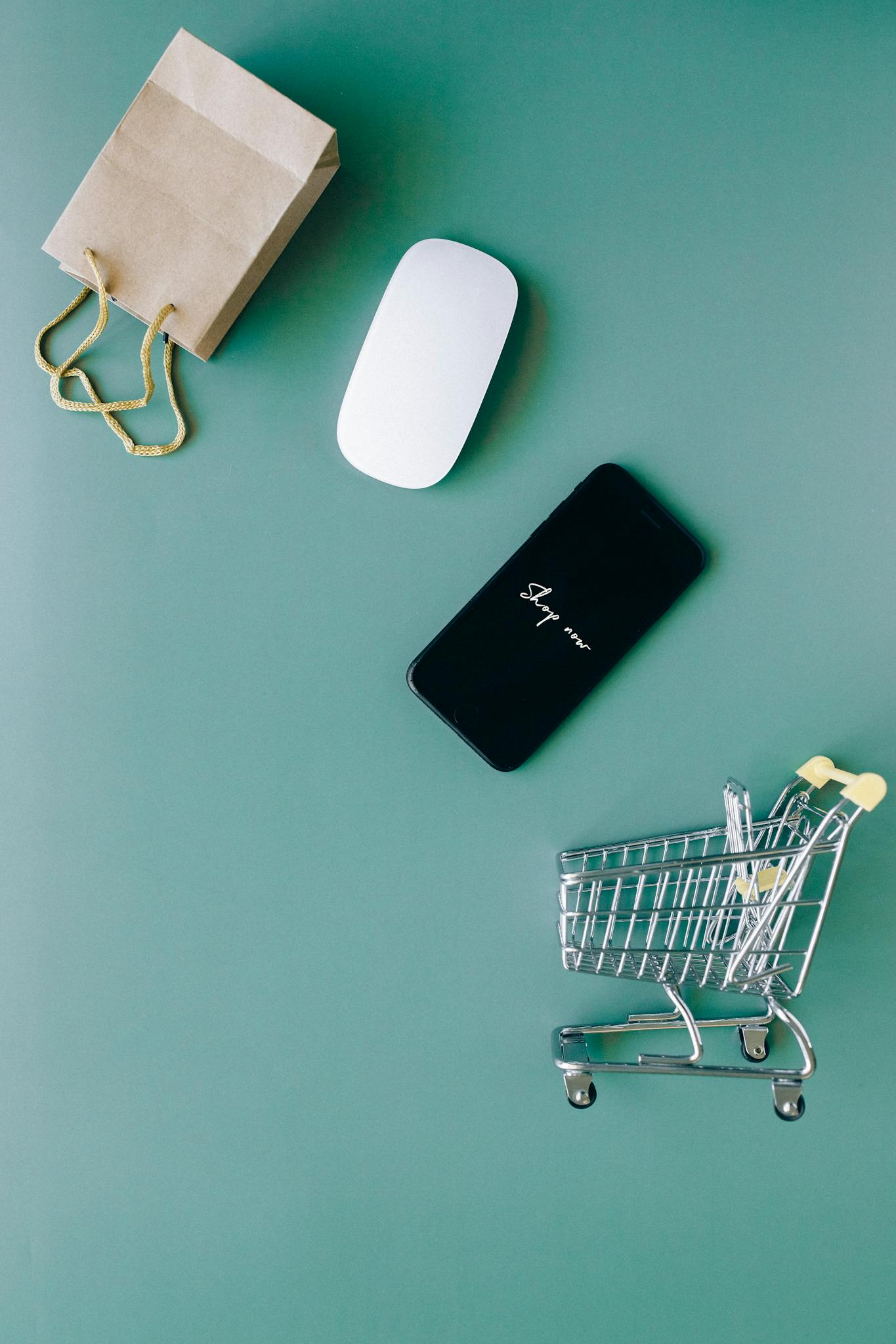 Top view of a smartphone, mouse, paper bag, and mini shopping cart on a green background, symbolizing e-commerce.